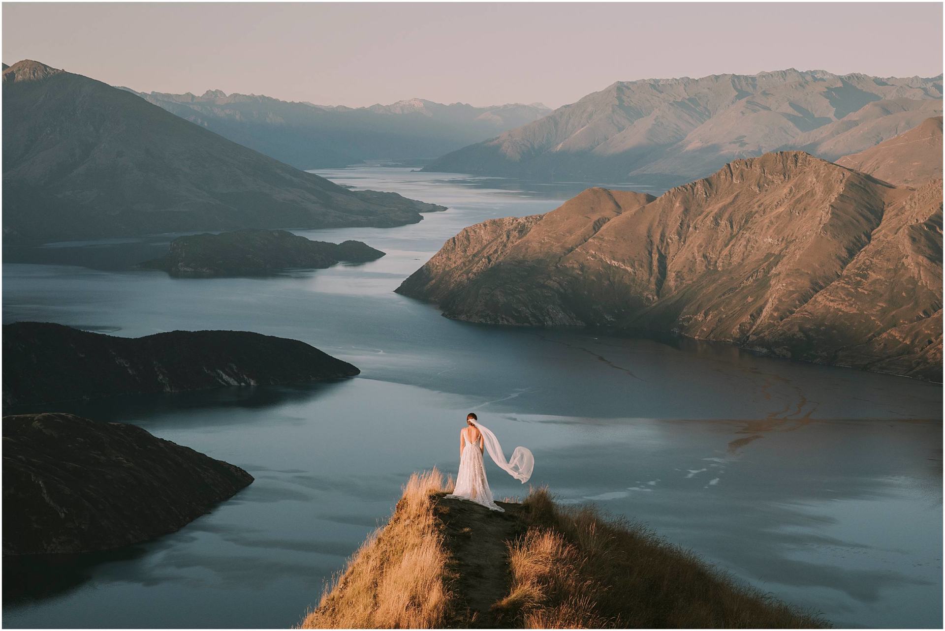 Charlotte Kiri Photography - Wedding Photography of a bride standing at the edge of Roy's Peak wearing a stunning low V-back dress, and long sheer veil that is floating romantically in the wind, as she looks over spectacular glacial lakes and mountains in Wanaka, New Zealand.