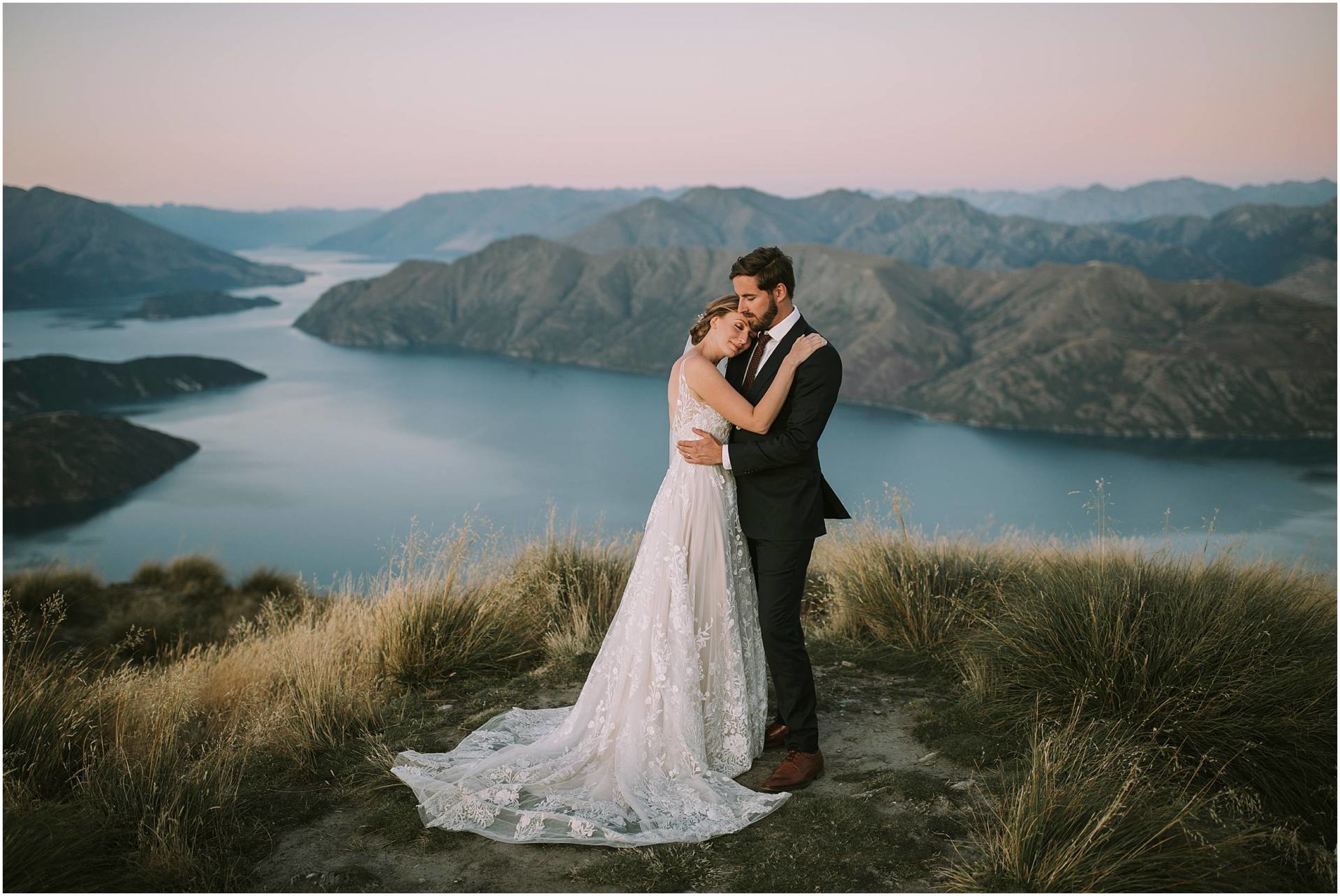Charlotte Kiri Photography - Wedding Photography of a bride wearing a pretty long sheer lace dress with a low back, hugging her groom who wears a smart black suit, as they stand on the edge of a peak overlooking glacial lakes and mountains in Wanaka, New Zealand.