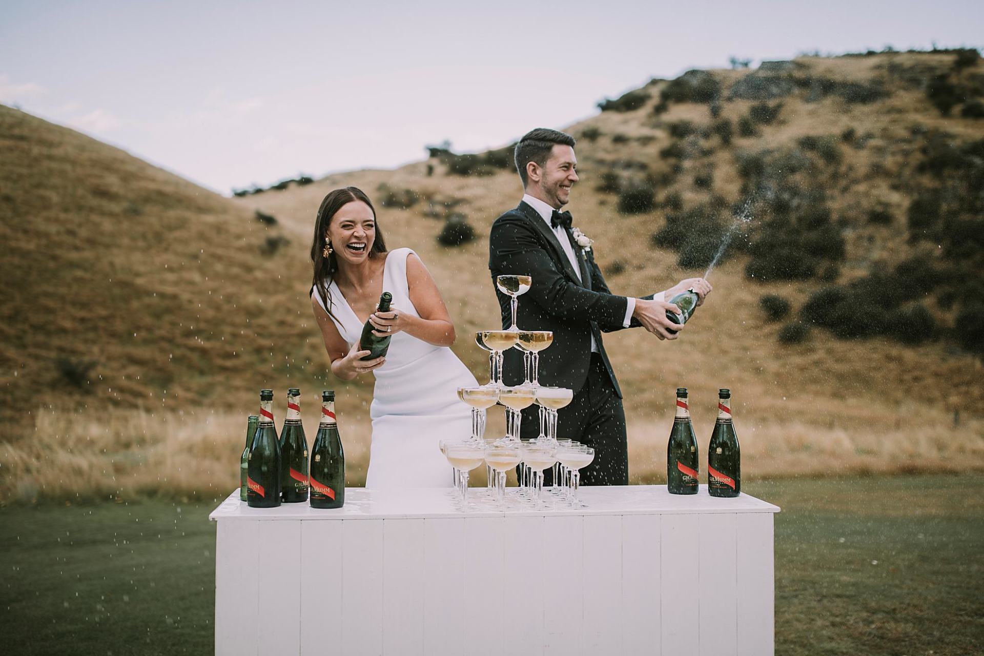 Charlotte Kiri Photography - Wedding Photography of a bride wearing an elegant fitted V-neck gown, and her groom in a black tuxedo with black bow tie, as they pop champagne corks, in front of a champagne glass pyramid in Wanaka, New Zealand.