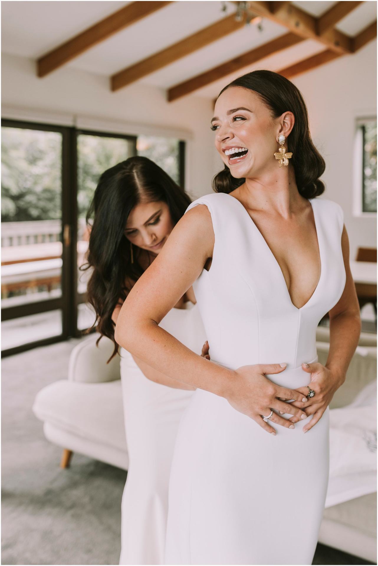 Charlotte Kiri Photography - Wedding Photography of a bride laughing as her bridesmaid helps button up her sleek low V-neck dress, before the wedding in their hotel room in Wanaka, New Zealand.
