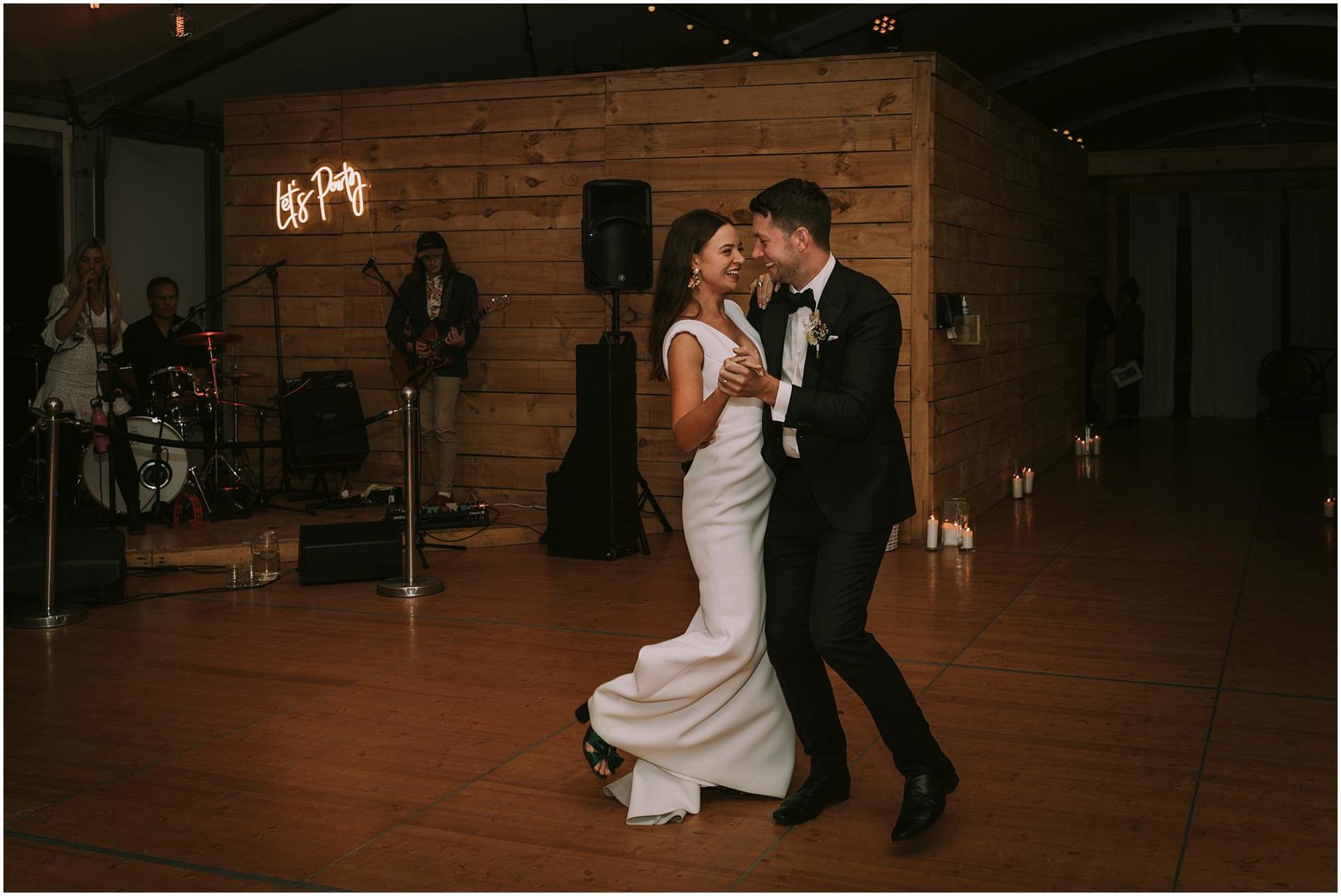 Charlotte Kiri Photography - Wedding Photography of a bride wearing an elegant fitted low V-necked dress as she dances with her groom in a black tuxedo, in front of a band at the wedding reception in Wanaka, New Zealand.