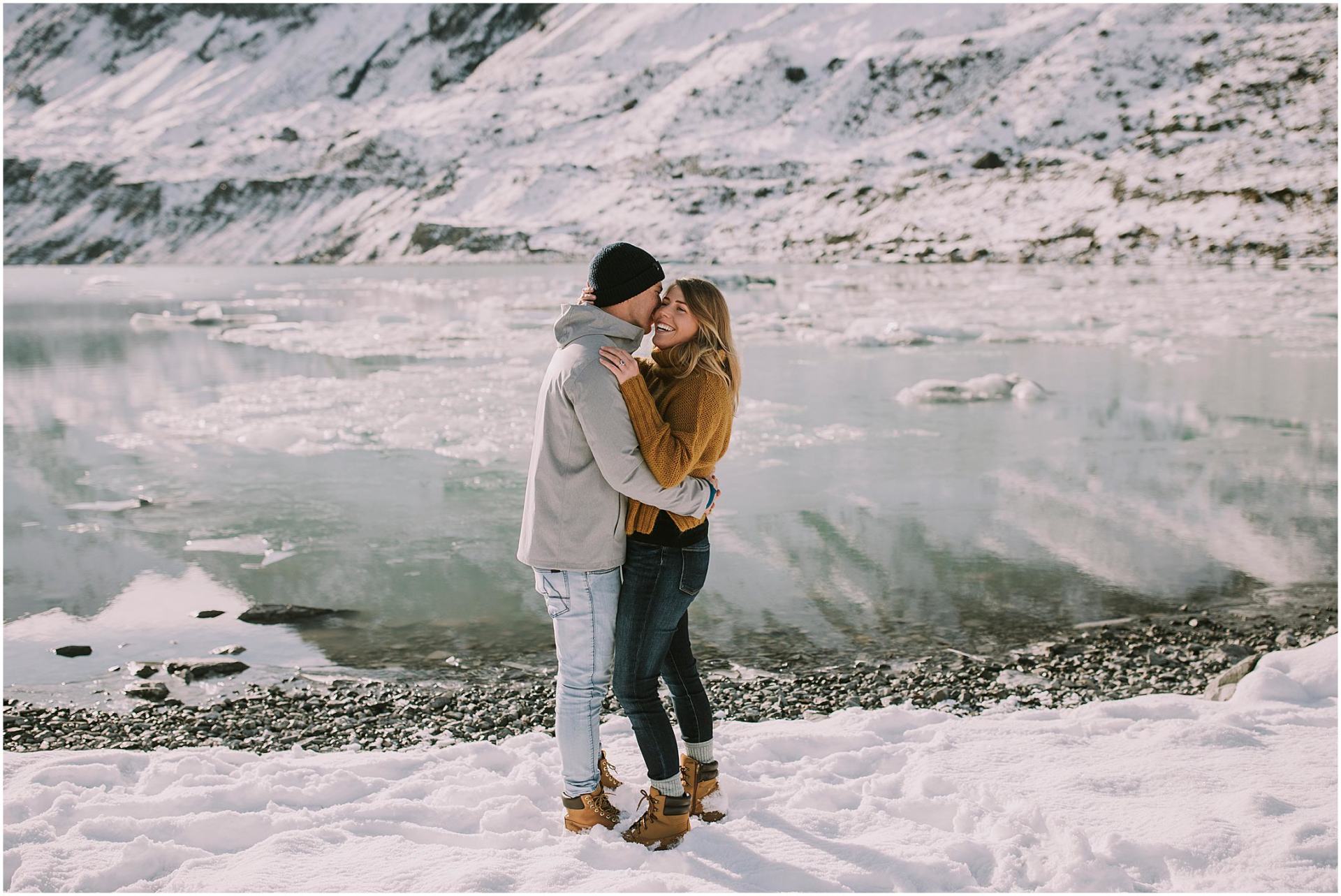 Charlotte Kiri Photography - Engagement Photography with the happy couple standing in the snow and embracing and smiling in front of a breathtaking glacial lake on Mount Cook, Aoraki, New Zealand