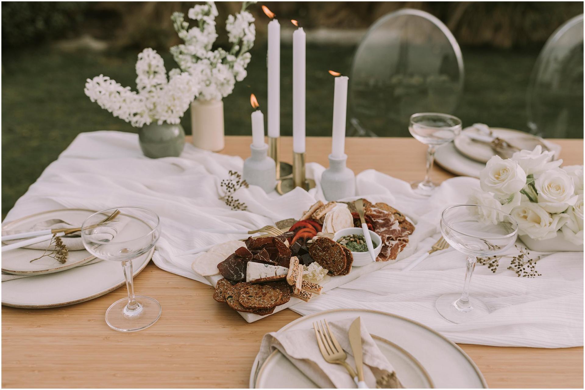 Charlotte Kiri Photography - Wedding Photography of a table setting with white floral arrangements and candles, a cheese board with cured meats, crackers, and place settings with champagne glasses and a damask-striped table runner in Wanaka, New Zealand.