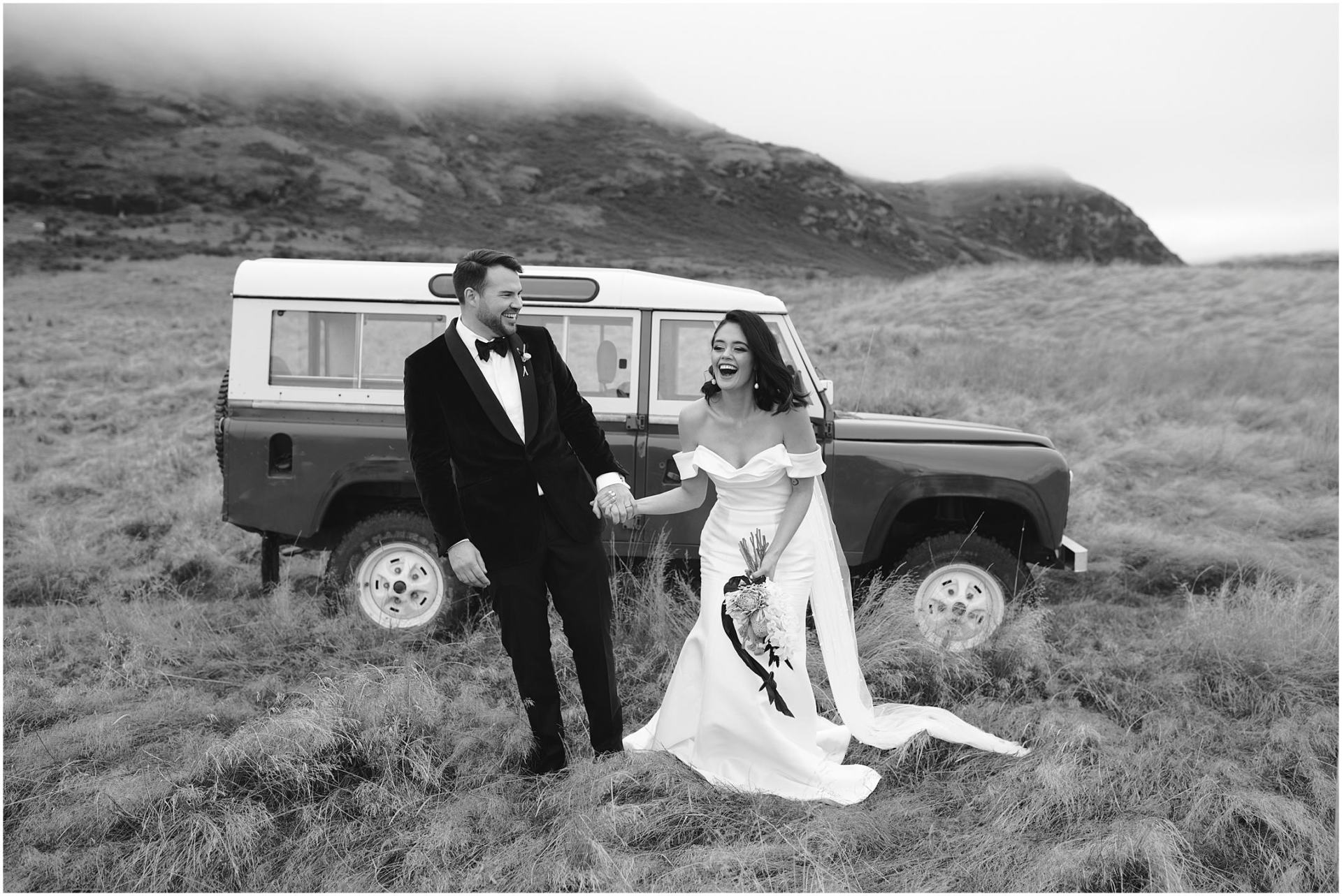 Charlotte Kiri Photography - Wedding Photography black and white of a bride wearing a gorgeous strapless dress holding hands, with her groom who wears a smart black tuxedo suit, as they walk hand in hand laughing in front of a Range Rover truck in a field with mountains behind in Wanaka, New Zealand.