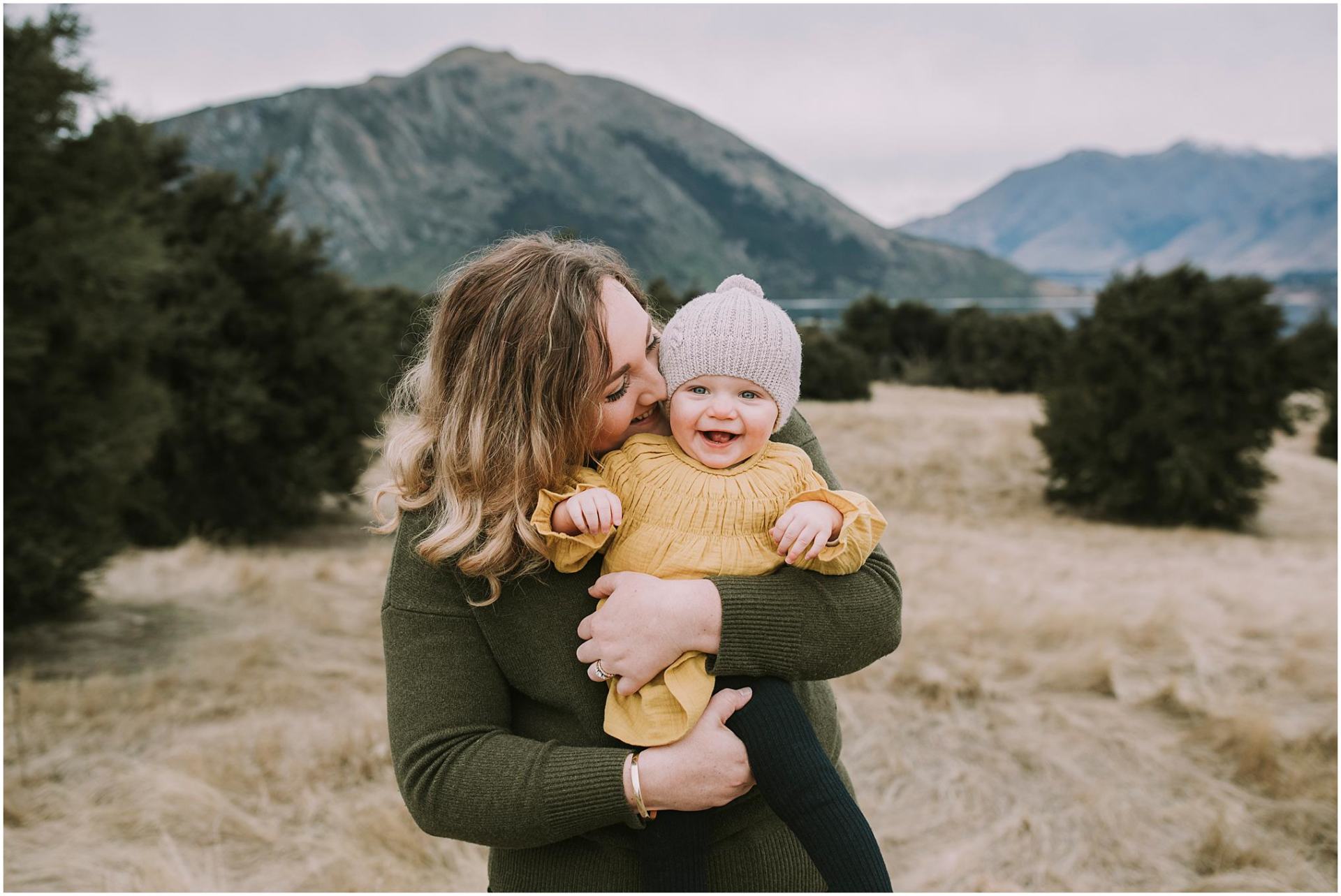 Charlotte Kiri Photography - Family Photography of a mother snuggling into her child as they laugh joyfully in her arms, in a field with mountains behind in Wanaka, New Zealand.