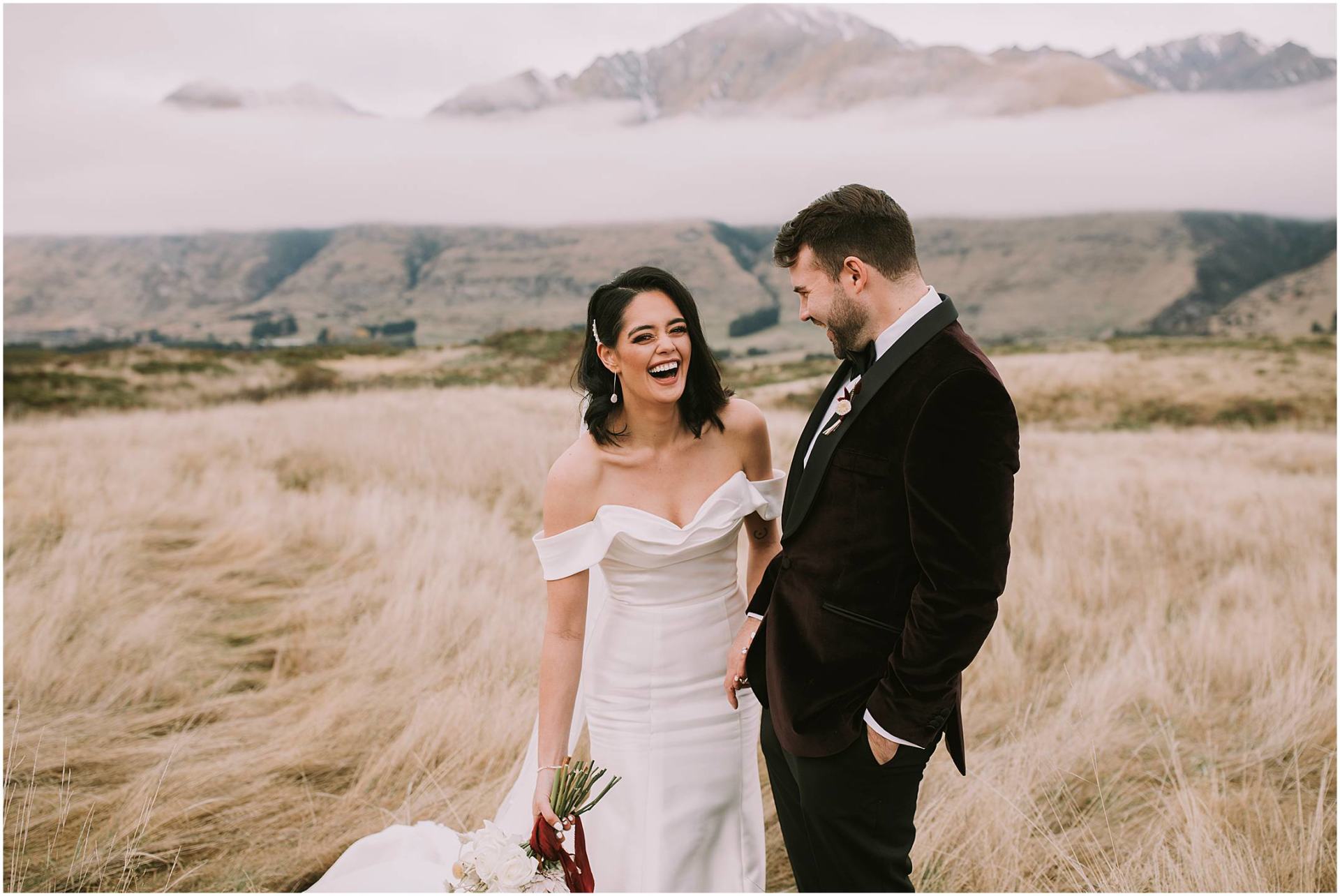 Charlotte Kiri Photography - Elopement Photography with bride laughing and wearing stunning off-shoulder gown with fitted skirt and long train, and groom wearing velvet dinner jacket and black pants, standing in the long grass with mountains and clouds behind in Wanaka, New Zealand