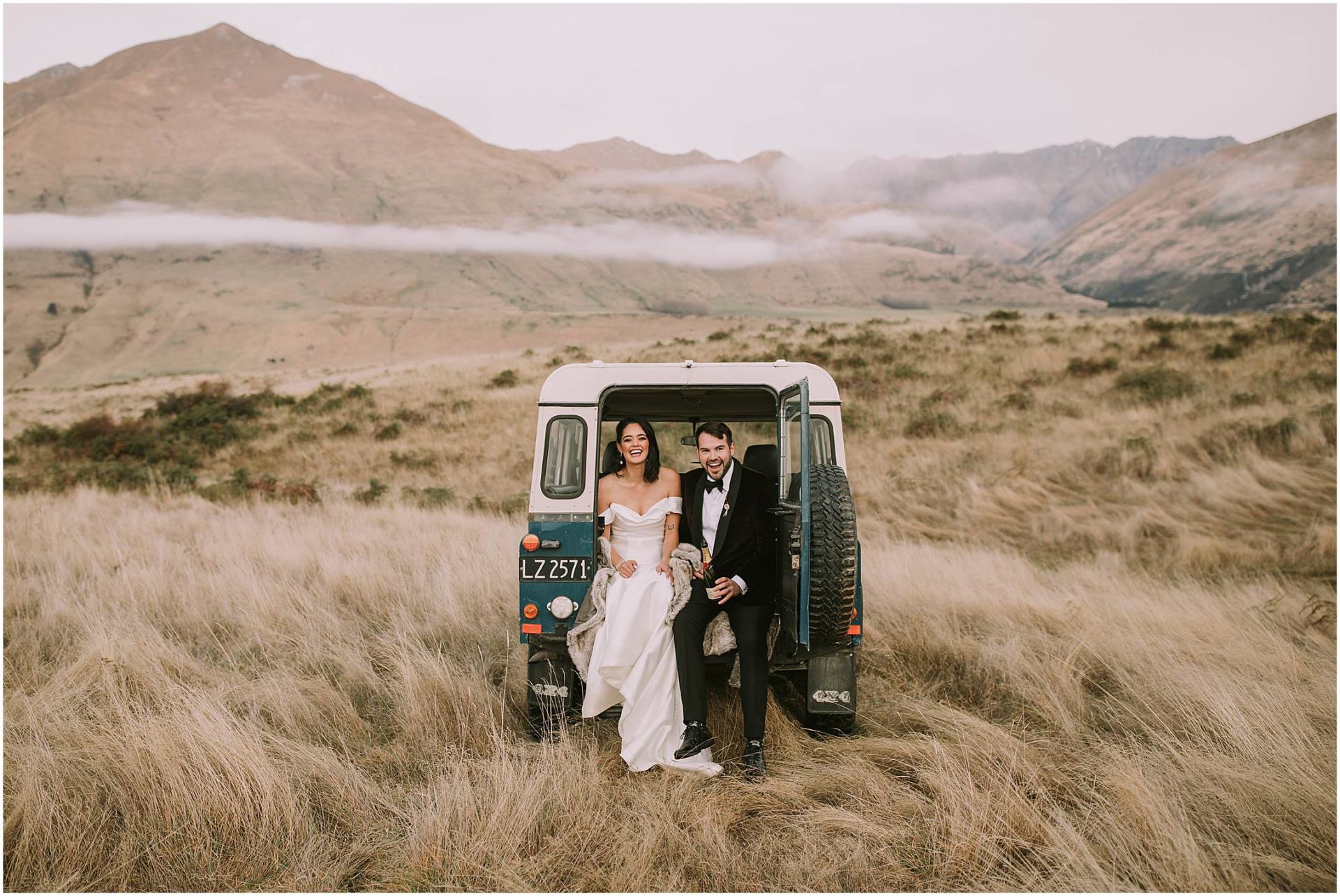 Charlotte Kiri Photography - Elopement Photography with the bride and groom sitting side by side laughing in the back of a Land Rover, that has been parked in a field of long grass in front of a backdrop of mountains and low hanging clouds in Wanaka, New Zealand