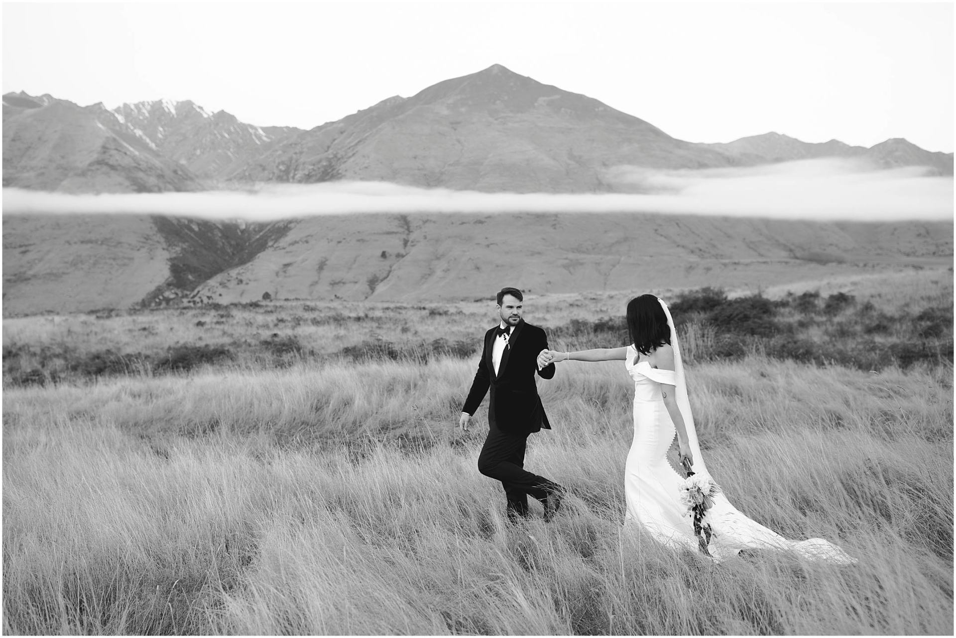 Charlotte Kiri Photography - Elopement Photography with bride wearing stunning off-shoulder gown and groom in a black dinner suit holding hands and walking through the long grass with a backdrop of mountains and long white clouds in Wanaka, New Zealand
