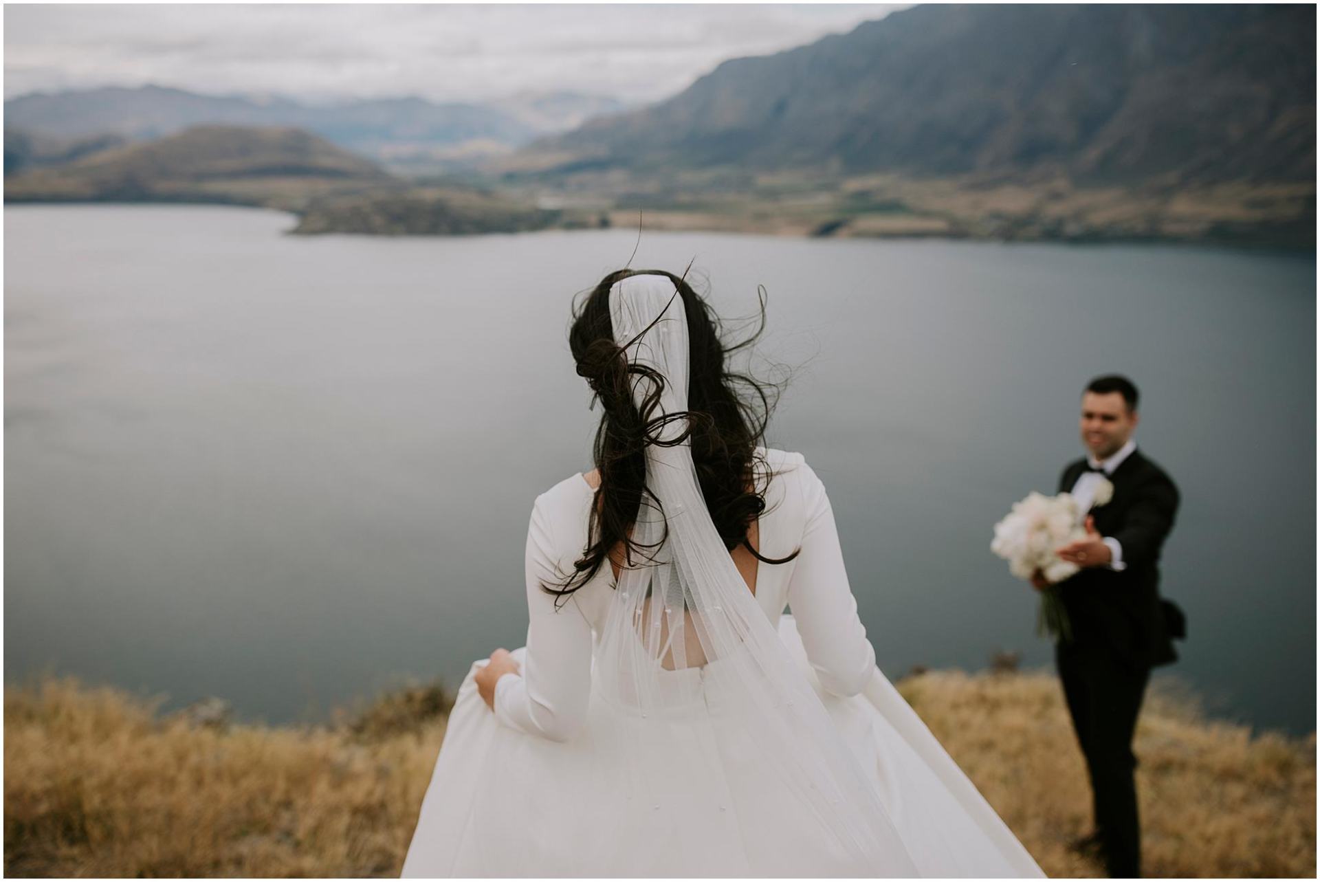 Charlotte Kiri Photography - Wedding Photography of a bride wearing an elegant low-back dress and sheer veil with pearls walking towards her groom as he holds her bouquet of white flowers and waits on the edge of a peak, with glacial lakes and mountains behind in Wanaka, New Zealand.