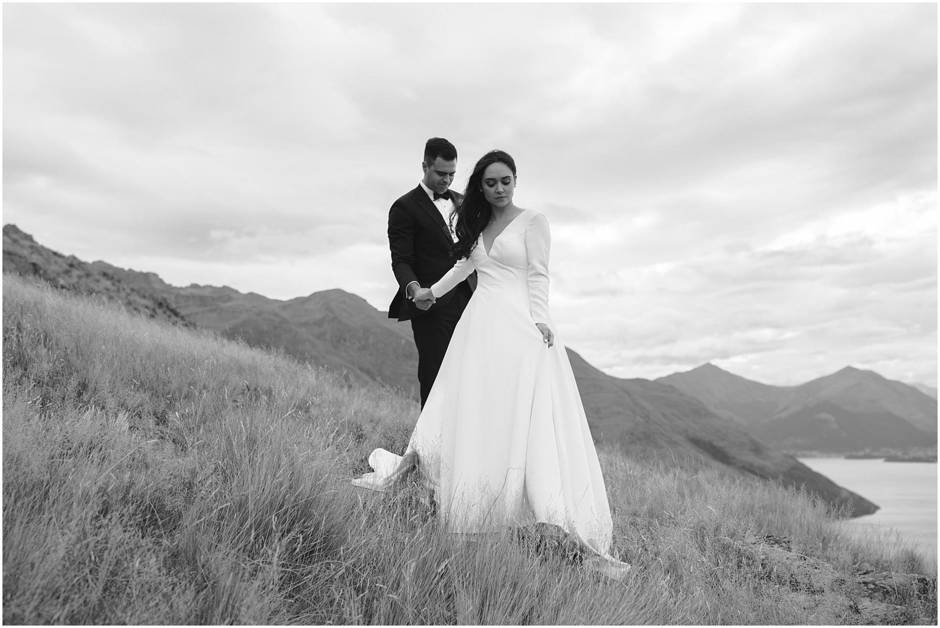 Charlotte Kiri Photography - Wedding Photography black and white image of a bride wearing an elegant V-necked long-sleeve dress with an A-line skirt, walking towards Coromandel Peak and holding hands with her groom who wears a smart black tuxedo suit in Wanaka, New Zealand.