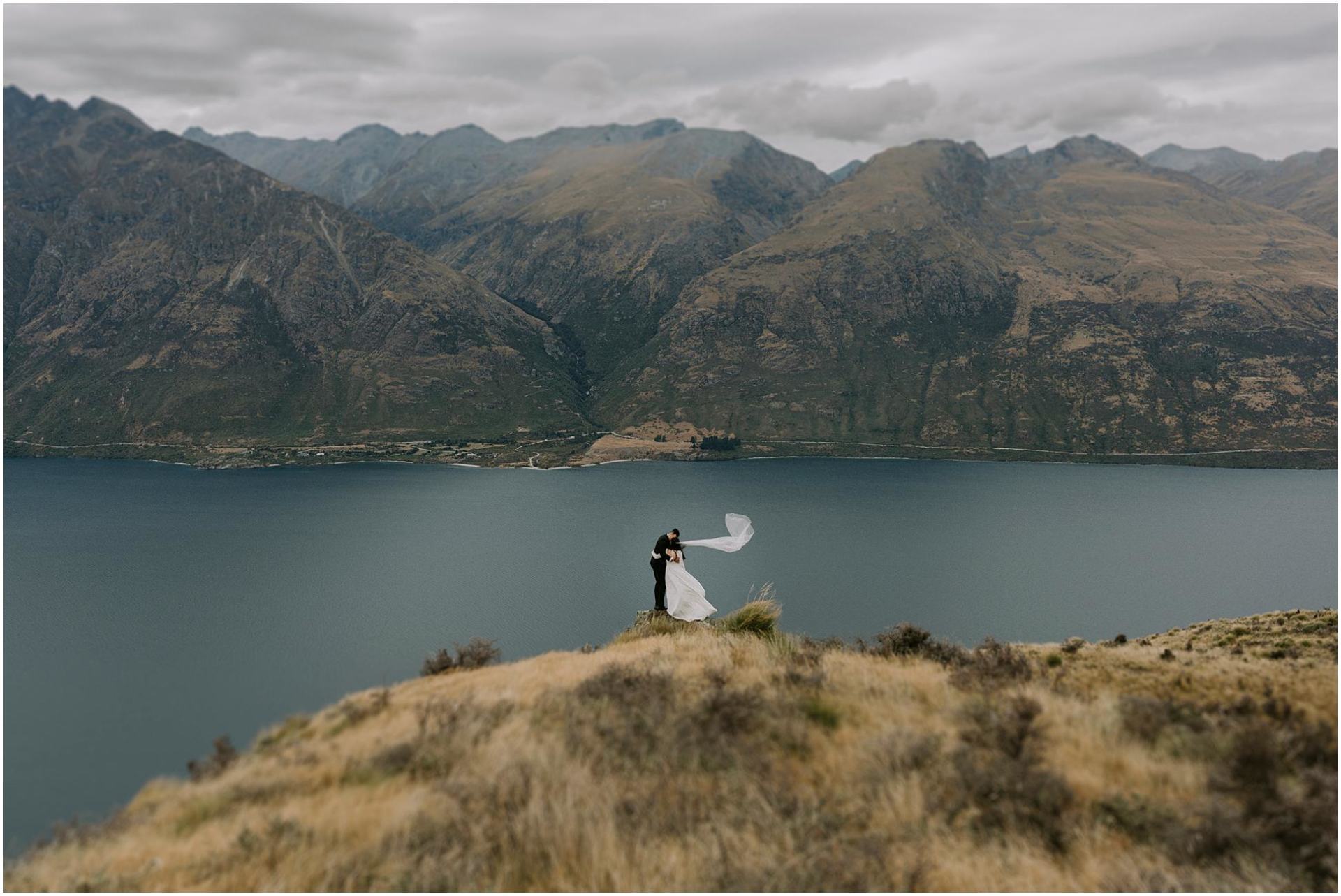 Charlotte Kiri Photography - Wedding Photography a breathtaking shot of a bride embracing her groom as her veil floats behind her on top of Coromandel Peak with an expanse of lakes and mountains behind in Wanaka, New Zealand.