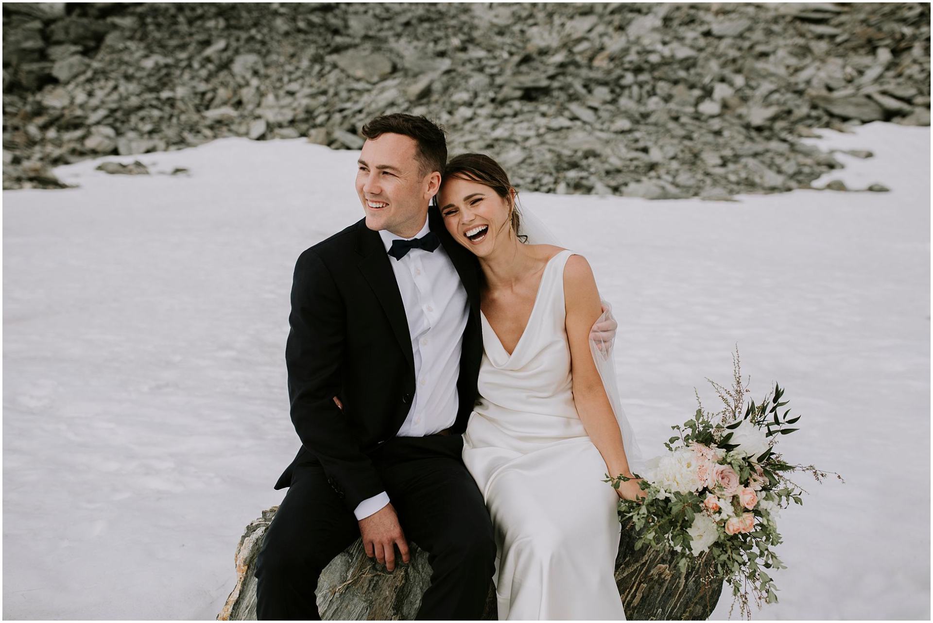 Charlotte Kiri Photography - Wedding Photography of a bride wearing a pretty satin cowl gown laughing and hugging her groom who is dressed in a black tuxedo as they sit together on a rock next to a glacier near Coromandel Peak in Wanaka, New Zealand.