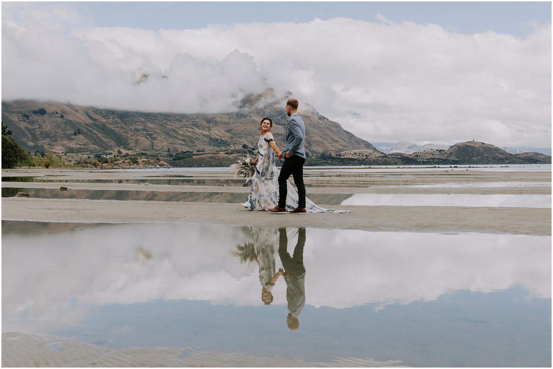 Charlotte Kiri Photography - Wedding Photography of a bride wearing a lovely blue floral dress with tasseled sleeves, holding hands and walking with her groom who wears a stylish grey jacket and black pants as they walk across a river bed near Coromandel Peak in Wanaka, New Zealand