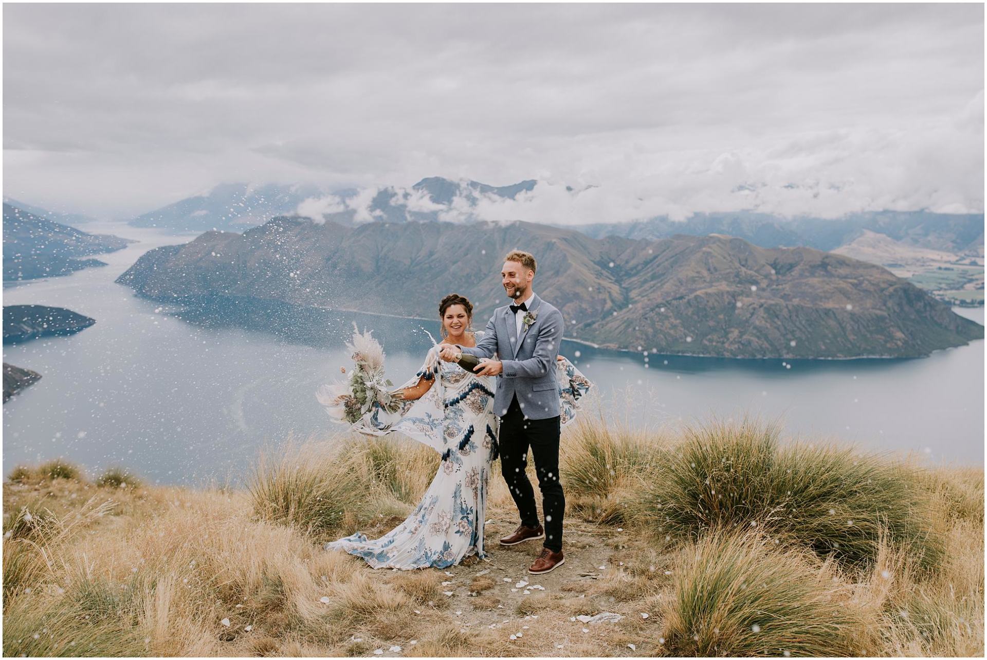 Charlotte Kiri Photography - Wedding Photography of a bride wearing a blue floral dress with tassels, and her groom wearing a grey jacket and black pants as they smile and open up a champagne bottle and spray the bubbles in the air in front of a peak overlooking lakes and mountains in Wanaka, New Zealand.