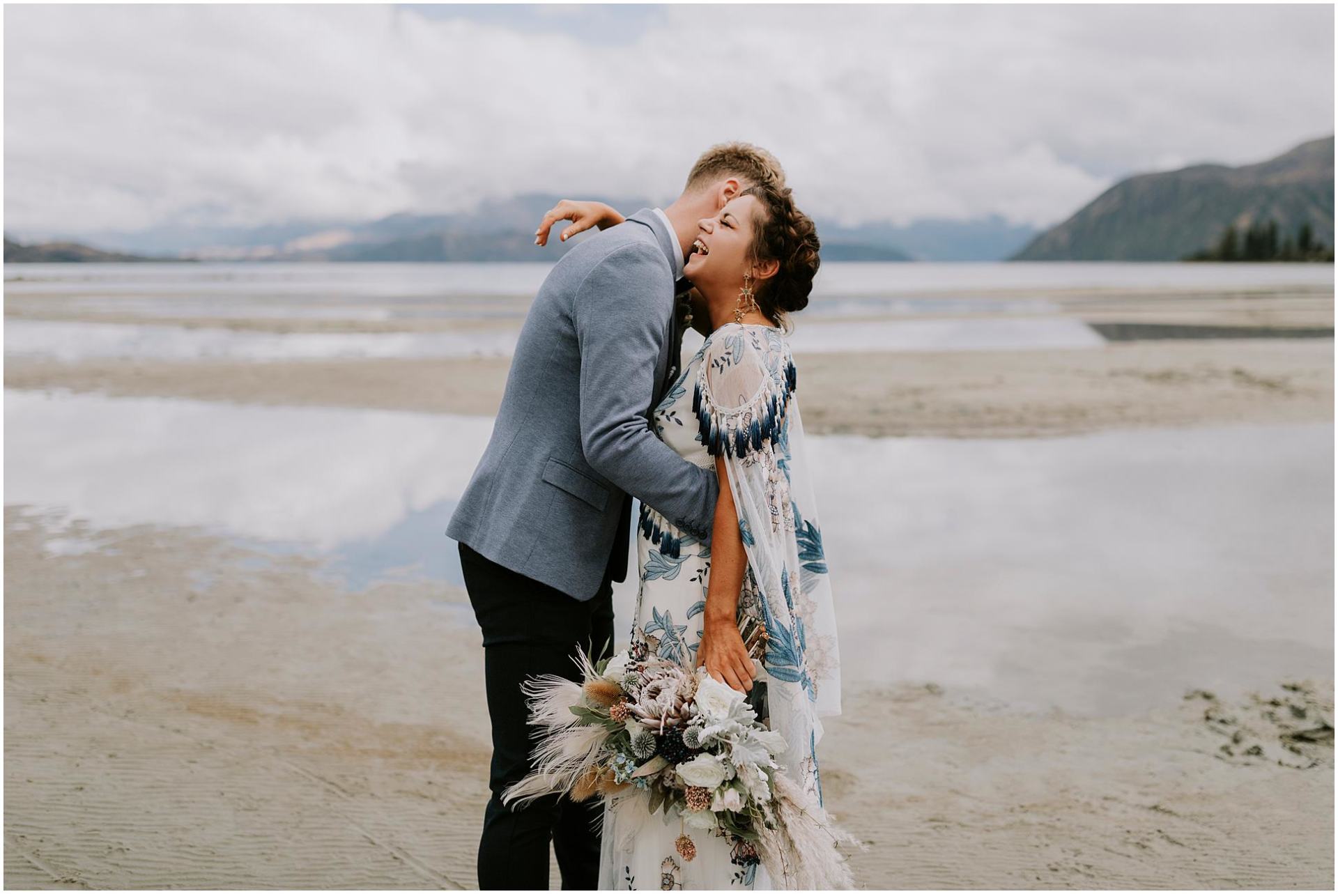 Charlotte Kiri Photography - Wedding Photography of a bride wearing a pretty blue floral dress with tassels, and her groom wearing a grey jacket and black pants as they embrace and laugh in front of lakes and mountains in Wanaka, New Zealand.