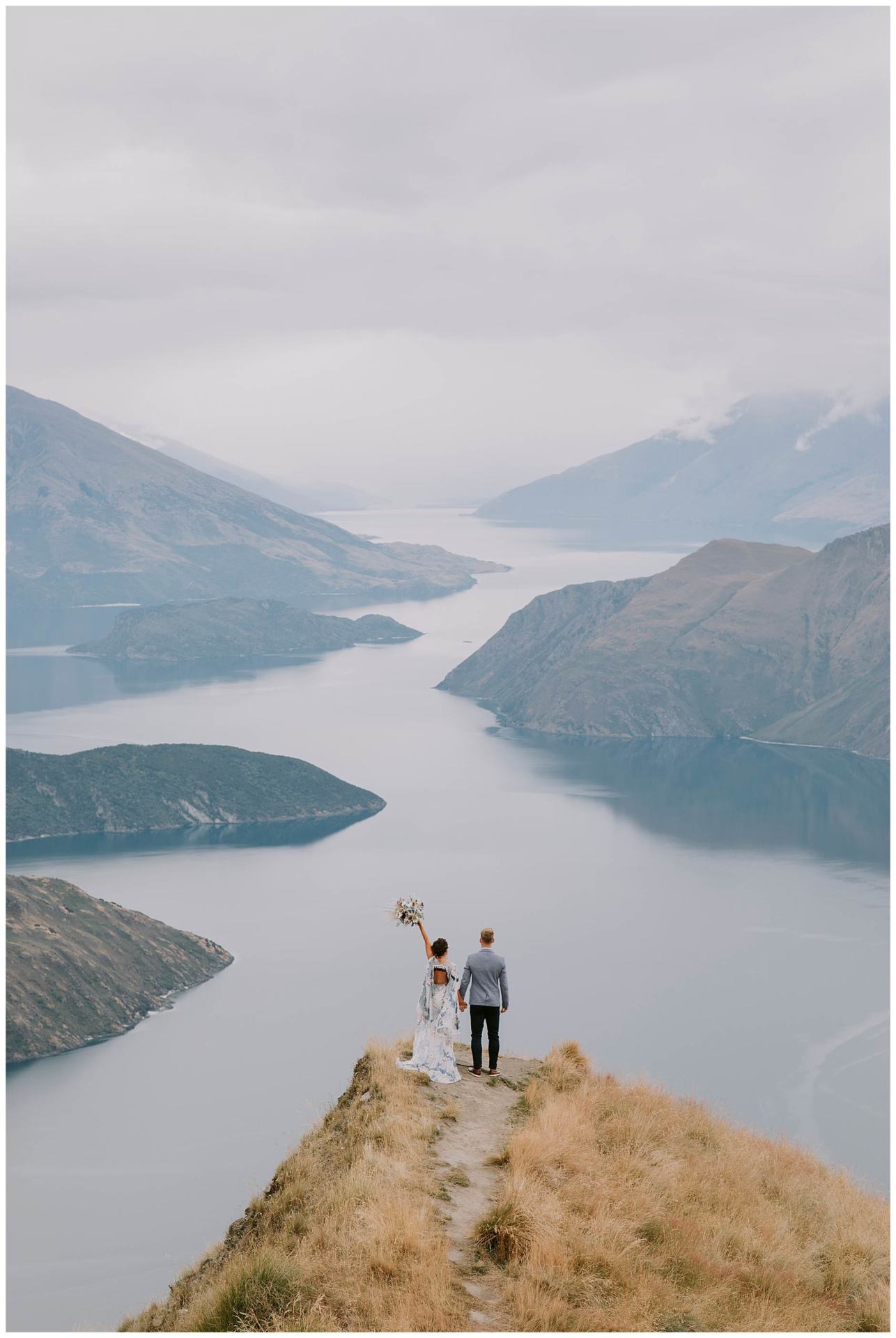 Charlotte Kiri Photography - Elopement Photography with the bride wearing a blue and white sheer floral dress and her bouquet is held high in the air, and the groom in a grey jacket and black pants, standing at the end Coromandel Peak with a backdrop of mountains and glacial lakes behind in Wanaka, New Zealand