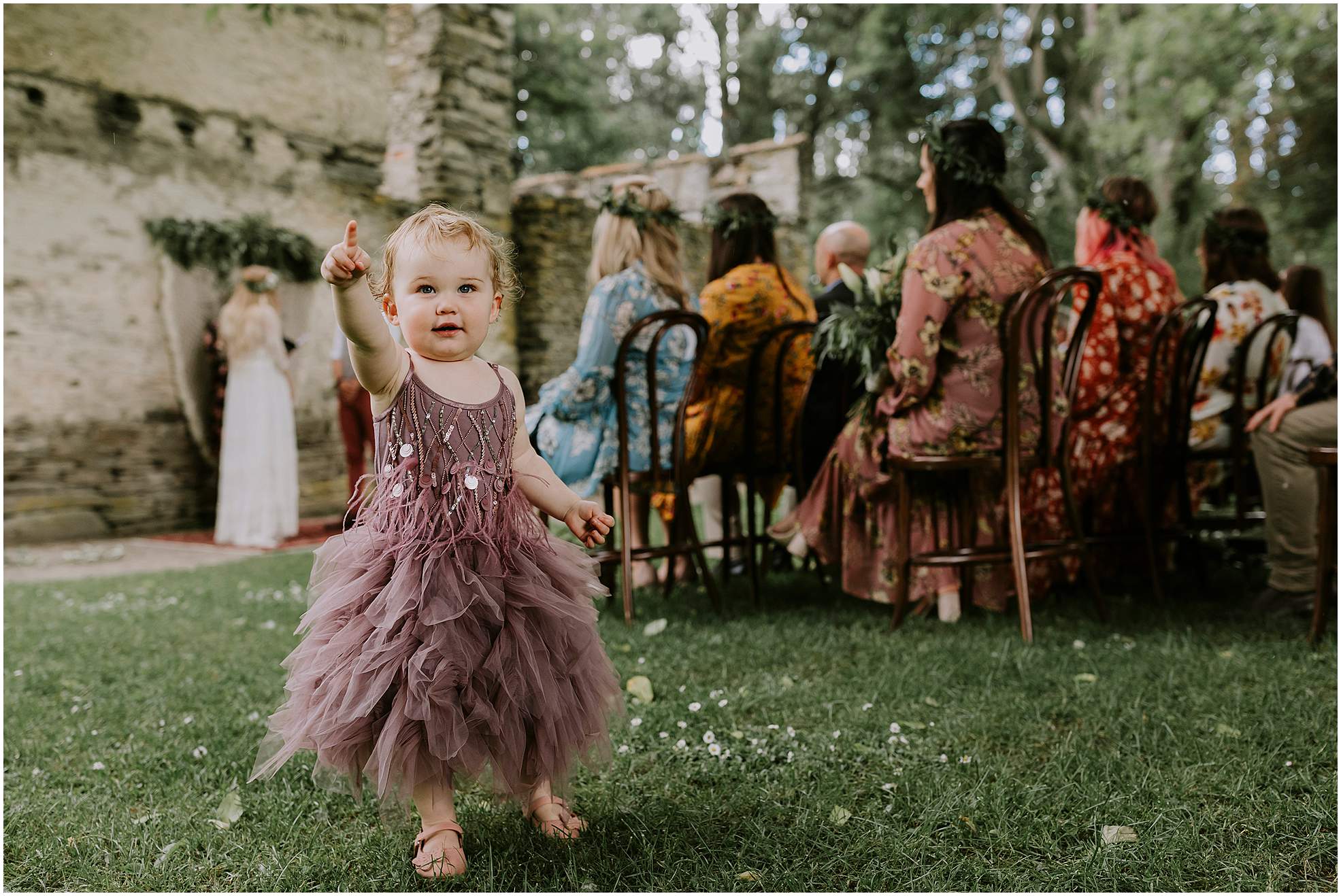 Charlotte Kiri Photography - Elopement photography with sweet little toddler wearing a dusty plum sequined dress and a tulle skirt, points up towards the photographer, whilst the wedding party who are dressed in long floral dresses with green wreaths in their hair, are seated at the alter listening to the vows of the bride and groom