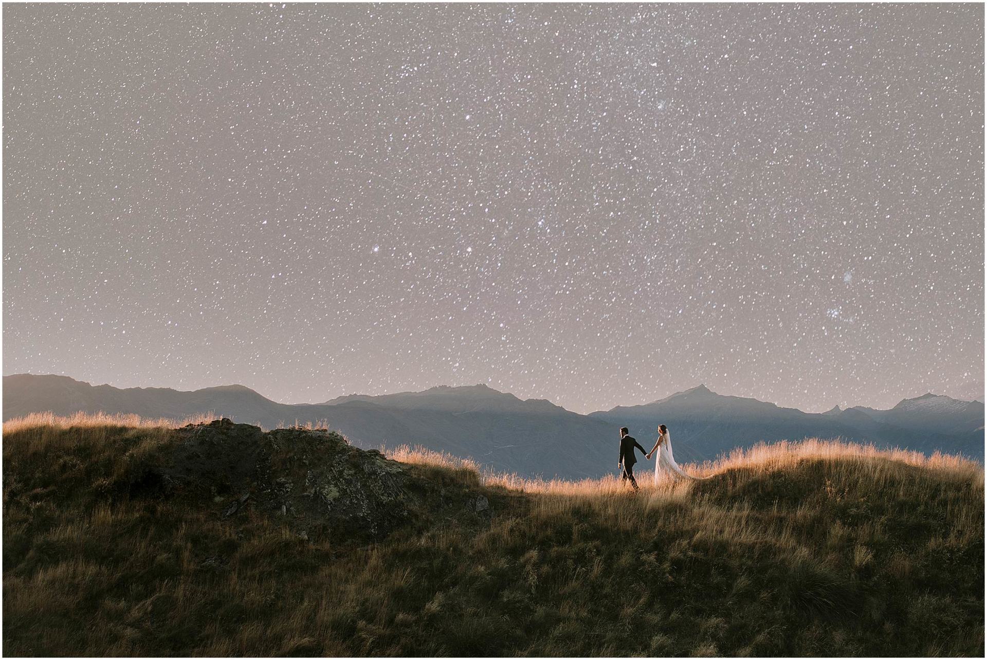 Charlotte Kiri Photography - Wedding Photography of a bride and groom holding hands and walking across undulating mountains with a backdrop of landscape and celestial sky behind them in Wanaka, New Zealand.