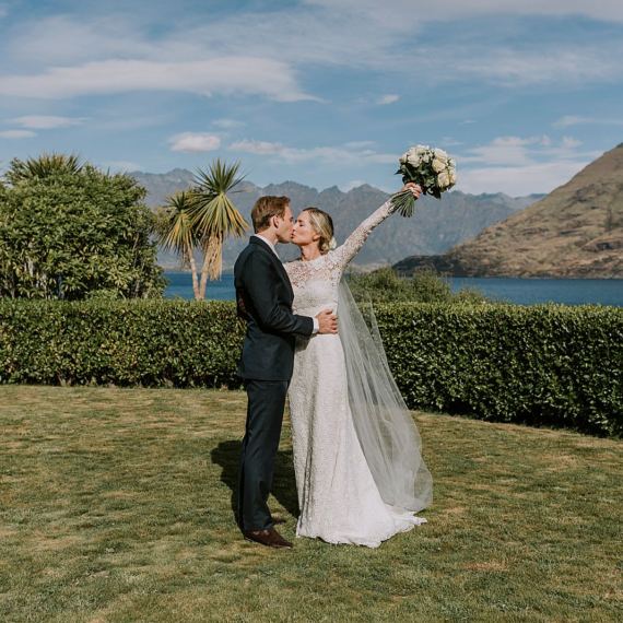 Charlotte Kiri Photography - Elopement Photography with bride wearing elegant long-sleeved lace gown holding her bouquet in the air as she kisses groom, who is wearing a black tuxedo suit, while they stand on hedge lined grass with the lake and mountains behind providing a stunning backdrop at Matakauri Lodge, Wanaka, New Zealand