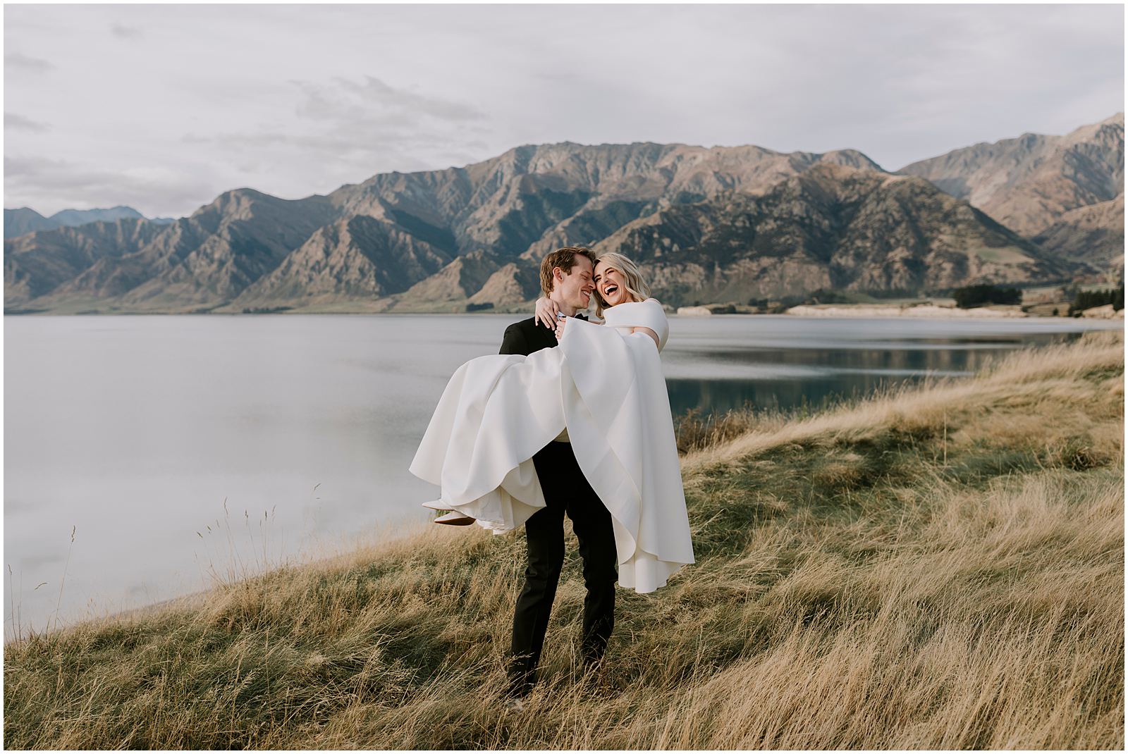 Charlotte Kiri Photography - Wedding Photography of a groom who wears a smart black suit, lifting his bride who wears a stunning white off-shoulder dress, as they stand on the edge of a picturesque glacial Lake with a backdrop of mountains in Wanaka, New Zealand.