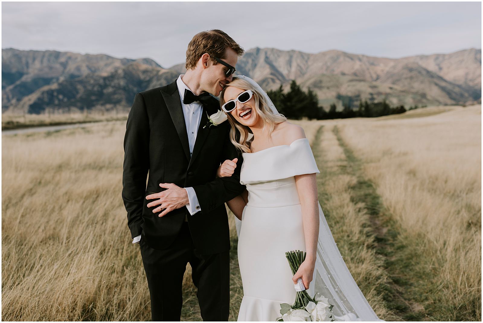 Charlotte Kiri Photography - Wedding Photography with a groom wearing a black tuxedo courteously escorting his wife who wears a lovely strapless gown and trendy white sunglasses, down a track in the grass of a big open field in Wanaka, New Zealand