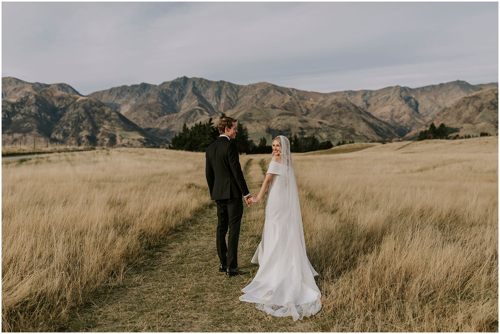 Charlotte Kiri Photography - Wedding Photography of a bride wearing a pretty off-shoulder gown with a long train and pearl embellished veil, holding hands with her groom who wears a smart black suit as they walk down a grass road in the middle of a large field, with a backdrop of mountains behind, in Wanaka, New Zealand.