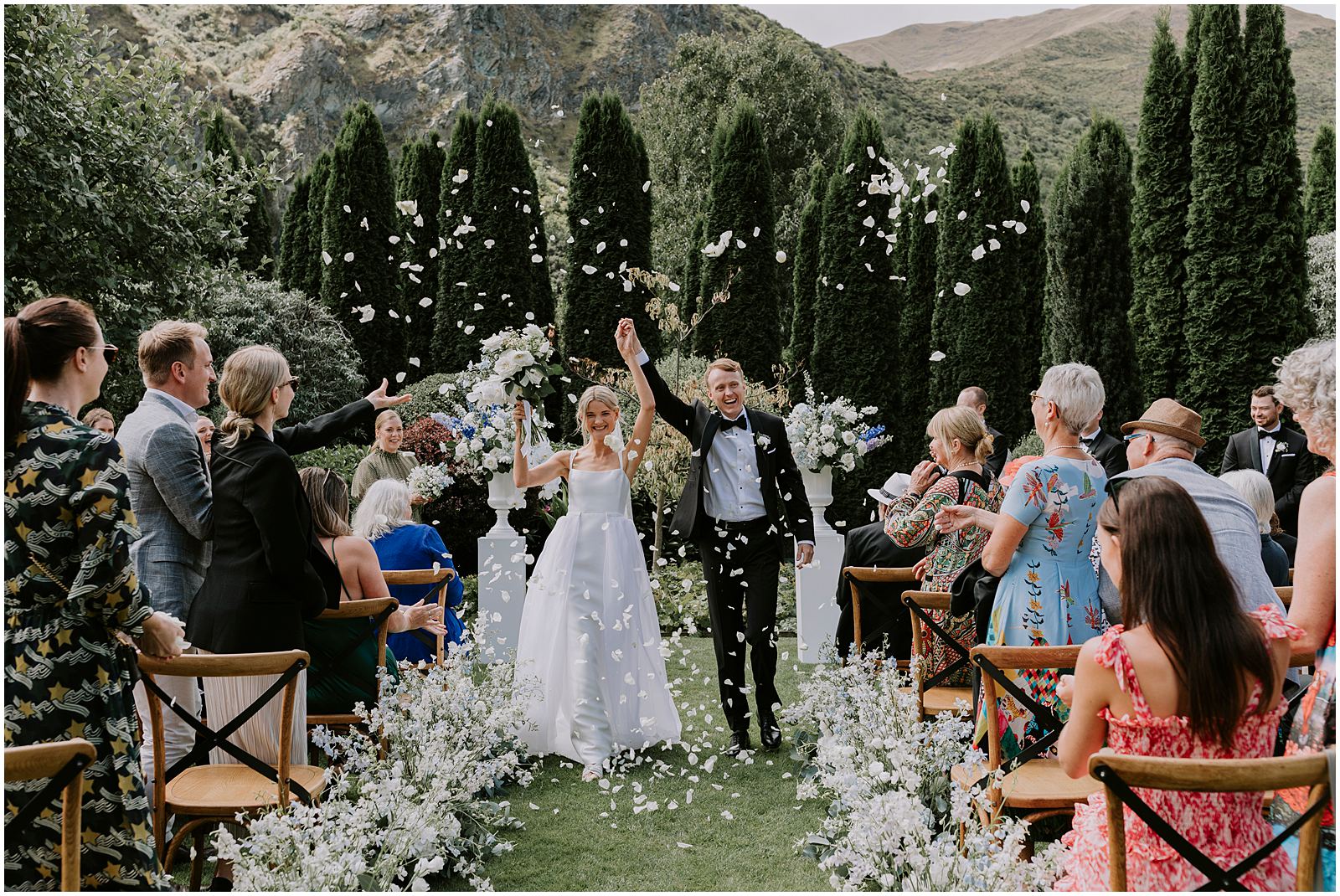 Charlotte Kiri Photography - Wedding Photography of a bride and groom walking down the aisle and raising their hands in the air together with joy, as they celebrate with their wedding guests who are throwing white petals around them, with a backdrop of poplar trees and mountains in Wanaka, New Zealand.
