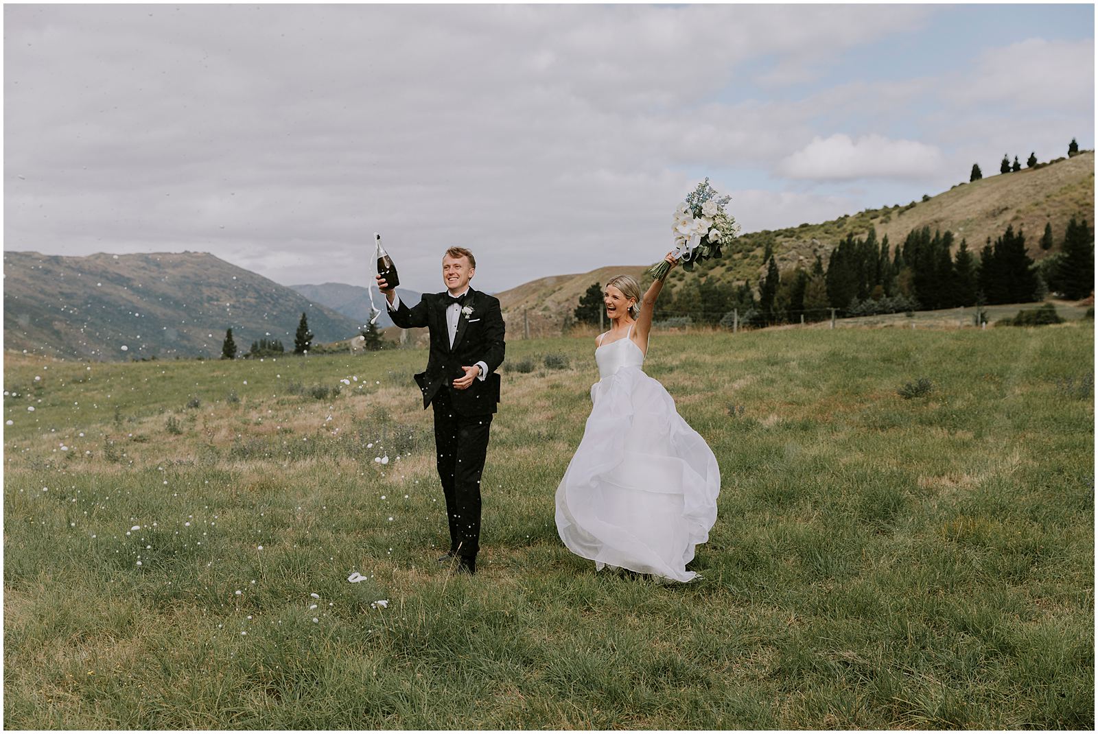 Charlotte Kiri Photography - Wedding Photography of a bride and groom walking in a field with a bottle of champagne spilling over as they raise their hands with joy, with a beautiful backdrop of mountains in Wanaka, New Zealand.