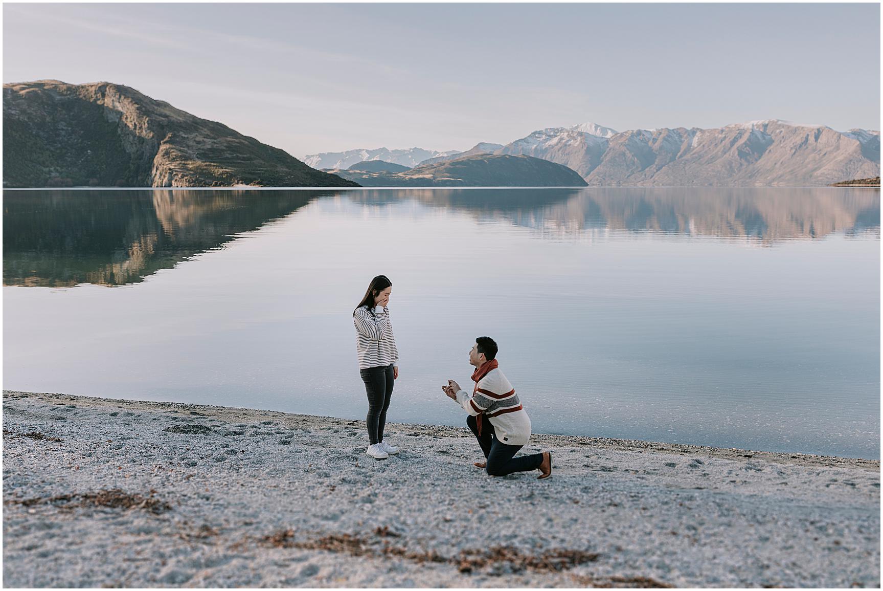 Charlotte Kiri Photography - Engagement Photography with a smart hopeful man proposing on bended knee to his happily stunned partner on the edge of a still lake with breathtaking snow-capped mountains behind at Matakauri Lodge in Wanaka, New Zealand