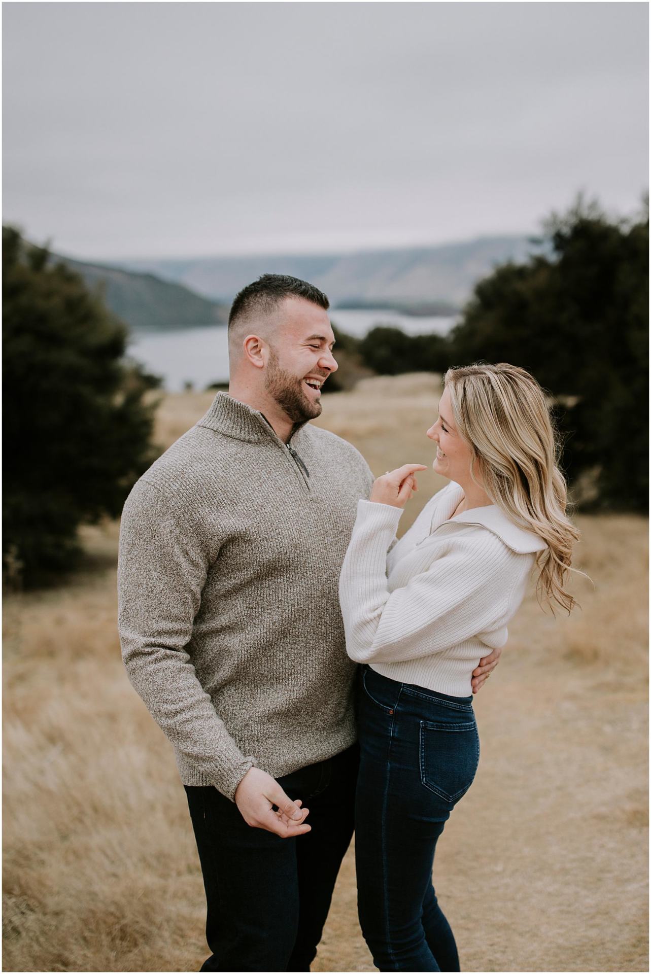 Charlotte Kiri Photography - Engagement Photography with a joyous couple embracing and laughing whilst admiring one another, in front of picturesque scenery of trees, lakes and mountains in Matakauri Lodge, Queenstown, New Zealand