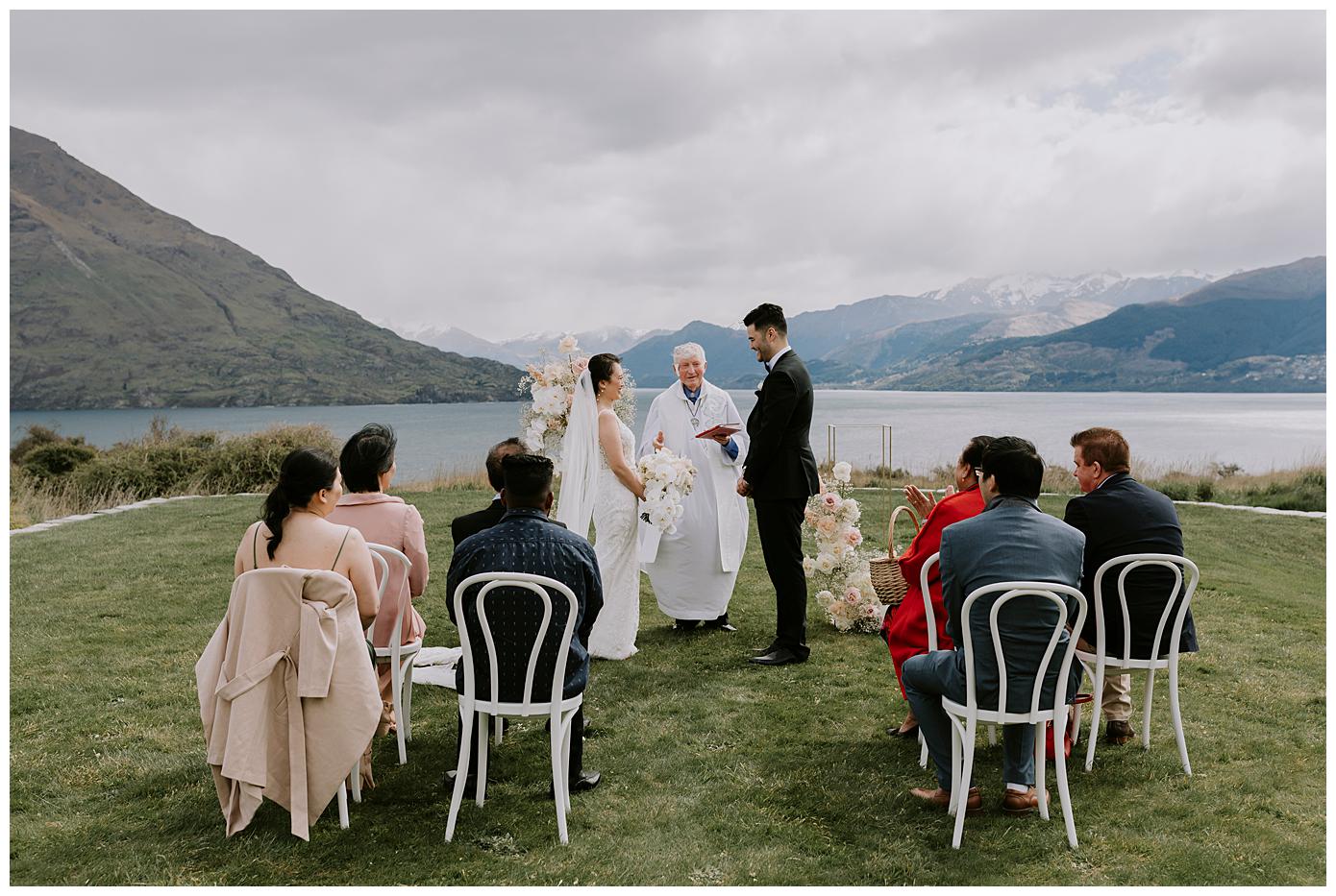 Charlotte Kiri Photography - Elopement Photography with the bride wearing a pretty long lace dress and sheer veil and the groom is wearing a black tuxedo suit, standing at the alter in front of a breathtaking backdrop of lakes and mountains at Jack's Retreat in Queenstown, New Zealand