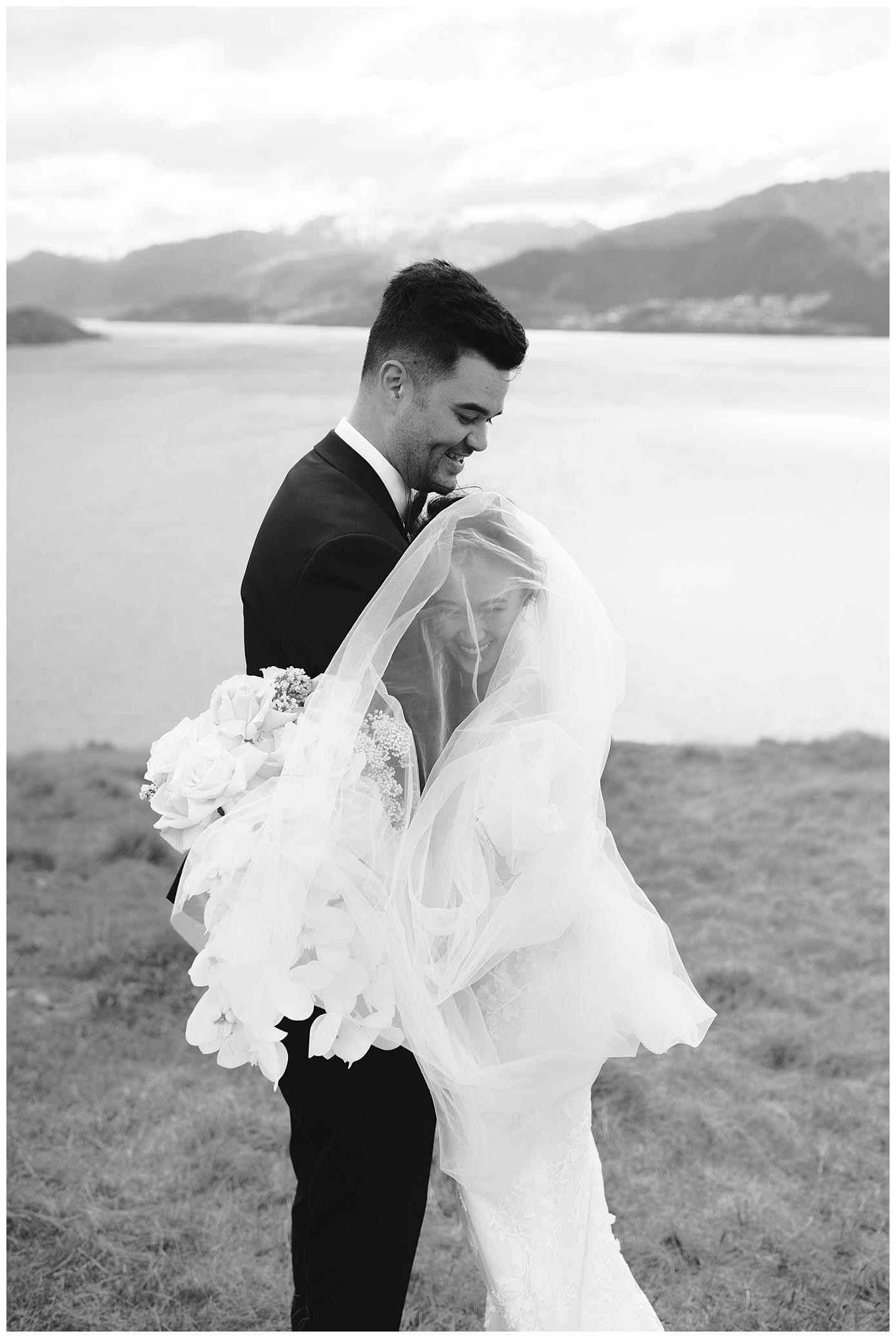 Charlotte Kiri Photography - Elopement Photography with a black and white picture of bride wearing long lace dress and hugging the groom, who wears a black tuxedo suit, as she nestles into his chest under her long sheer veil whilst holding a pretty bouquet of white flowers, with a stunning backdrop of lakes and mountains behind at Jack's Retreat in Queenstown, New Zealand