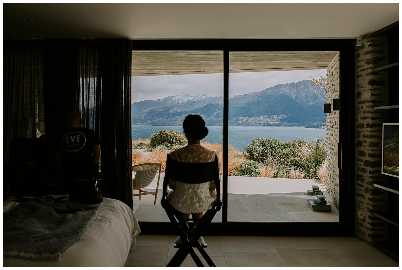 Charlotte Kiri Photography - Elopement Photography with bride wearing pretty floral lace gown sitting in a directors chair awaiting her makeup artist, whilst admiring the stunning view of lakes and mountains out the window, at Jack's Retreat in Queenstown, New Zealand