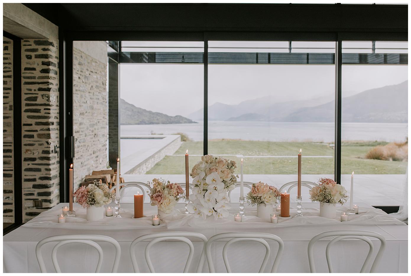 Charlotte Kiri Photography- Elopement Photography with an elegant table setting adorned with pretty white and blush floral centre pieces, copper candles, white mini tea light candles and a crisp white tablecloth, set in front of an epic view of the lakes and mountains at Jack's retreat in Queenstown New Zealand