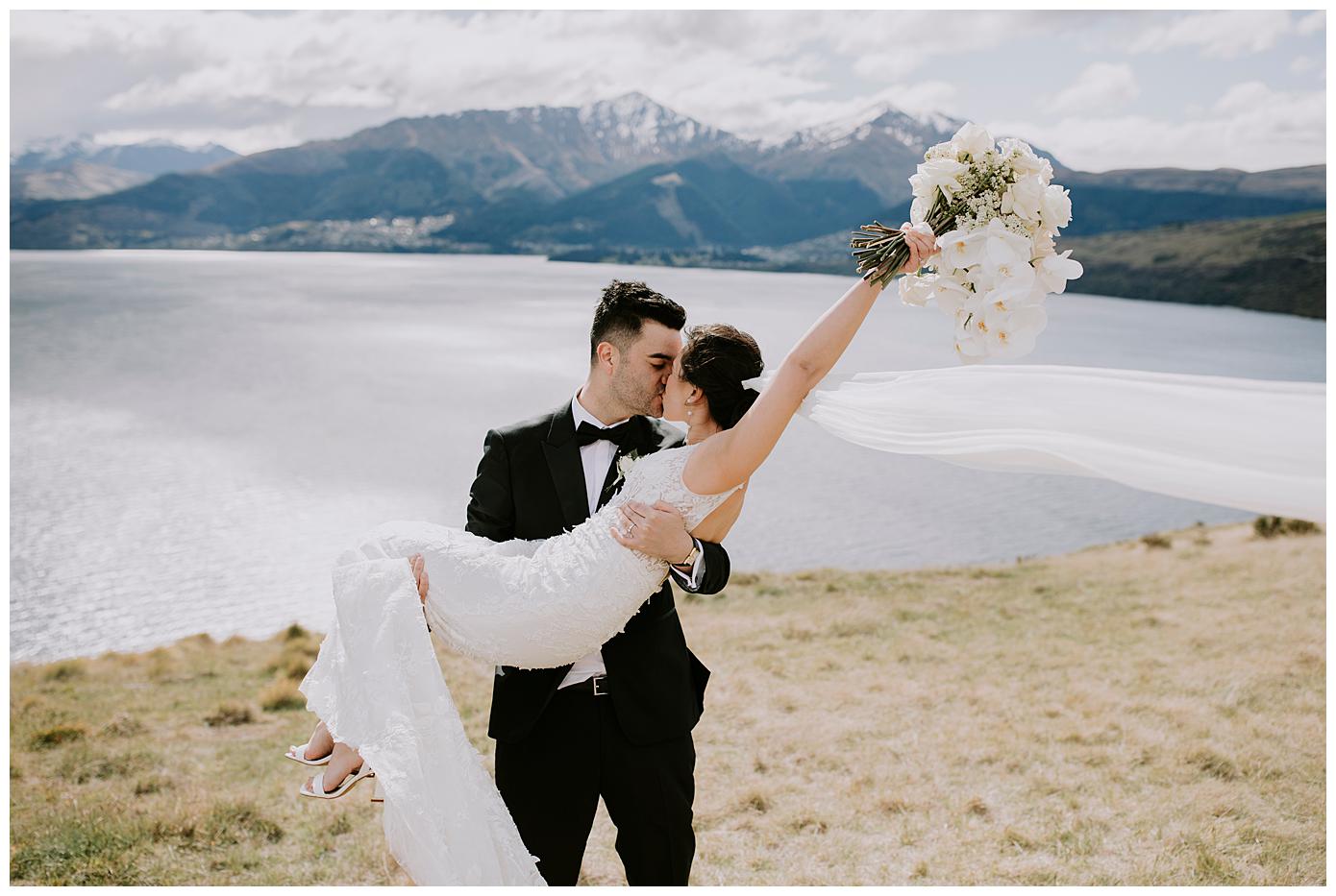 Charlotte Kiri Photography - Elopement Photography of groom wearing black tuxedo suit who is lifting up his bride who is wearing a lovely lace dress with long sheer veil that is floating in the wind, in front of a pictureque backdrop of lakes and mountains at Jack's retreat in Queenstown, New Zealand