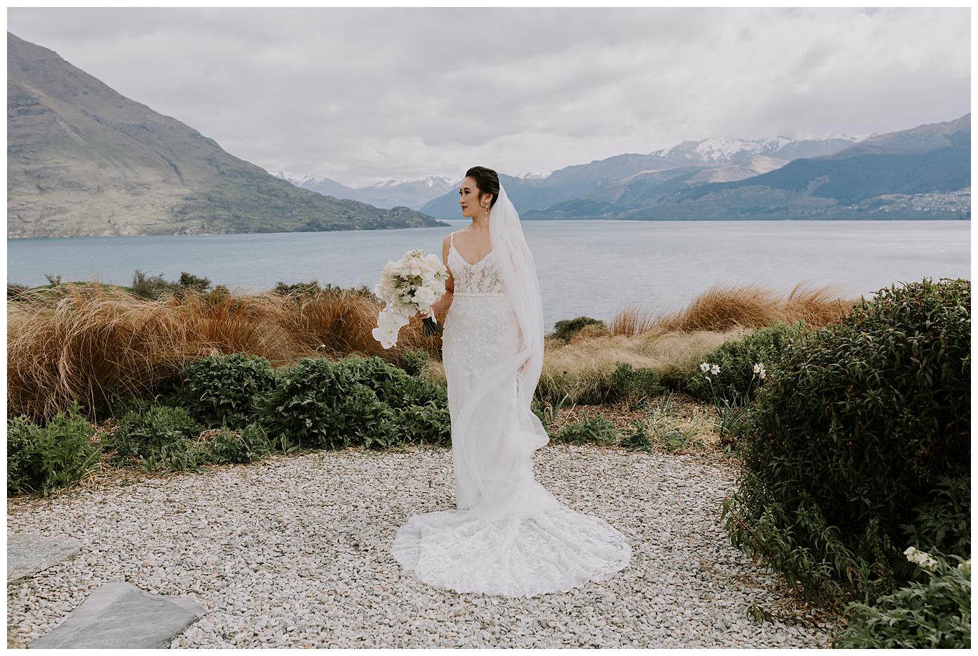 Charlotte Kiri Photography - Elopement Photography with bride wearing gorgeous floral lace gown with long train and sheer veil, holding pink and white bouquet in front of a stunning backdrop of lakes and mountains at Jack's Retreat in Queenstown, New Zealand